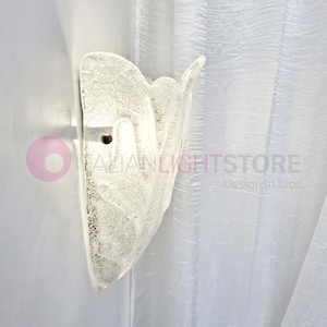 May include: A textured, frosted glass wall sconce with a unique, organic shape. The sconce is mounted on a white wall, and the design includes a small, circular opening for a light source. The text "ITALIANLIGHTSTORE" is visible.