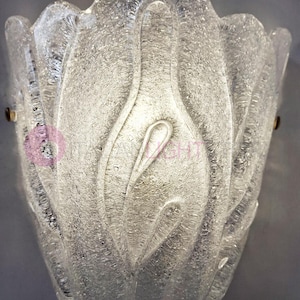 May include: A clear, textured glass wall sconce with a floral design. The sconce has a textured surface and is mounted with two small brass-colored screws. The overall shape resembles a stylized flower or bud.