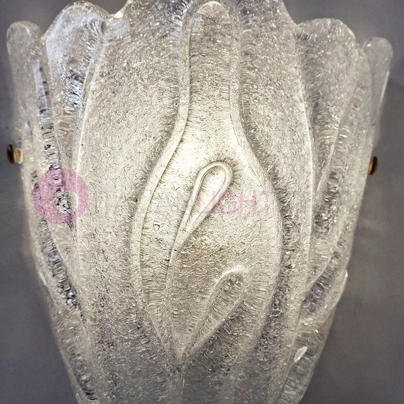 May include: A clear, textured glass wall sconce with a floral design. The sconce has a textured surface and is mounted with two small brass-colored screws. The overall shape resembles a stylized flower or bud.