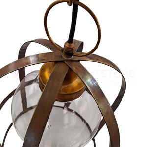 May include: A vintage-style pendant light fixture with a clear glass globe encased in a bronze-colored metal frame. The fixture has a circular top ring and a black cord. The metal has a distressed finish.