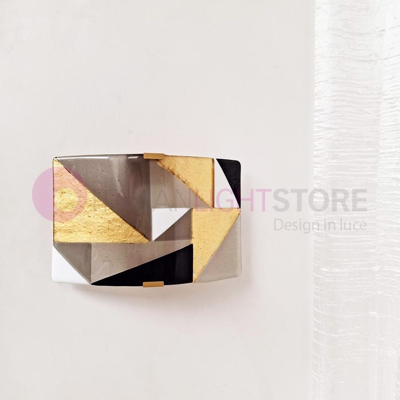 K&ouml;nnte beinhalten: Geometrische Wandkunst mit rechteckiger Form, die goldene, schwarze, wei&szlig;e und graue Dreiecke enth&auml;lt. Das Design umfasst goldene Akzente und eine moderne &Auml;sthetik. Das Kunstwerk ist an einer wei&szlig;en Wand befestigt.