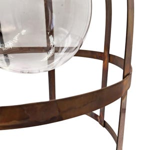 May include: A clear glass globe vase sits within a rusted metal cage. The cage has a circular base and vertical bars that support the globe. The metal has a weathered, aged appearance. The vase is empty and transparent.