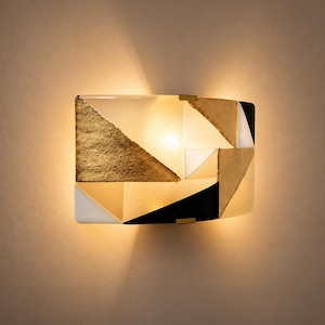 K&ouml;nnte beinhalten: Geometrische Wandleuchte mit goldenem, schwarzem, wei&szlig;em und beigem Design. Die Leuchte hat eine rechteckige Form mit gew&ouml;lbter Front und wird von innen beleuchtet, wodurch ein warmes Licht entsteht.