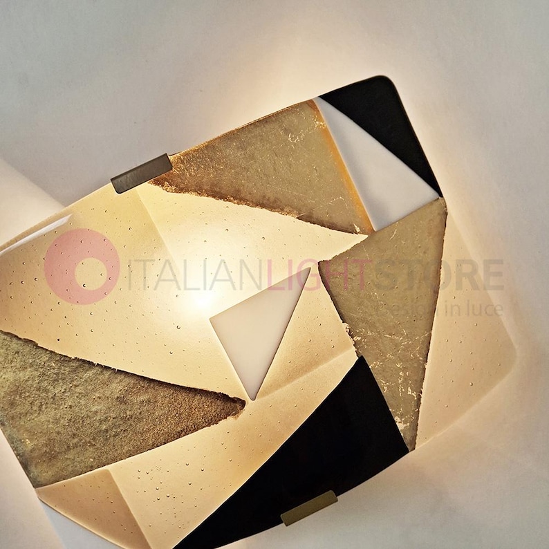 K&ouml;nnte beinhalten: Geometrische Wandleuchte mit einem geschichteten Design. Die Leuchte weist eine Mischung aus beige, gold, schwarzen und wei&szlig;en Dreiecksformen auf. Die Leuchte hat ein warmes Licht, das von der Mitte ausgeht und mit zwei kleinen Metallhalterungen befestigt ist.