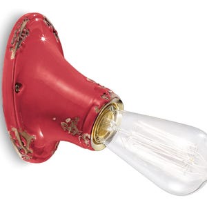 Puede incluir: Una lámpara de pared de cerámica roja con un acabado desgastado y una bombilla de vidrio transparente. La lámpara tiene una forma redondeada y una base de metal dorado.