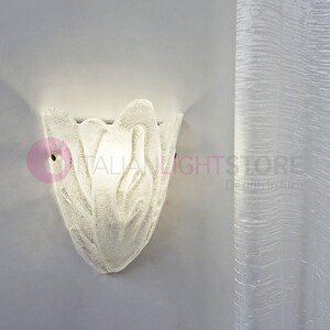 May include: A frosted glass wall sconce in a floral design. The light fixture is mounted on a light gray wall. The sconce has a textured surface and a small hole for mounting. The text "ITALIAN LIGHTSTORE" is partially visible.