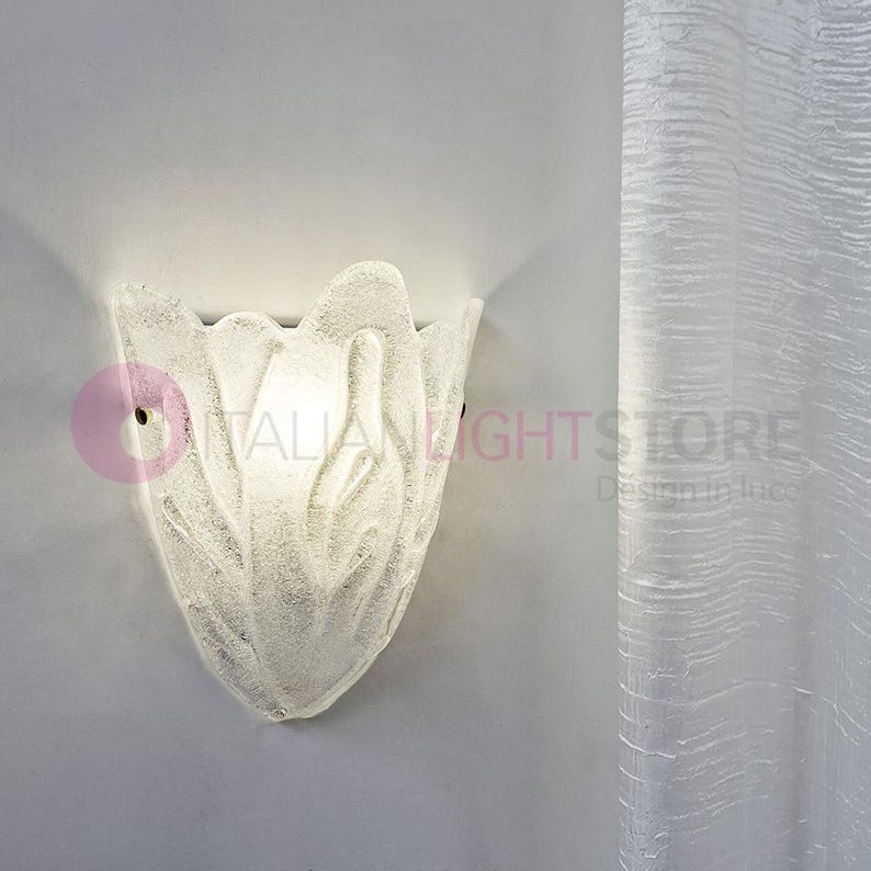 May include: A frosted glass wall sconce in a floral design. The light fixture is mounted on a light gray wall. The sconce has a textured surface and a small hole for mounting. The text "ITALIAN LIGHTSTORE" is partially visible.