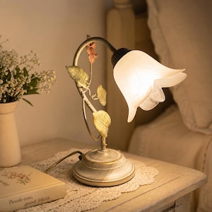 Puede incluir: Lámpara de noche decorativa con diseño floral. La lámpara presenta una pantalla blanca en forma de tulipán y una base con un acabado envejecido y antiguo. El tallo de la lámpara está adornado con hojas doradas y una pequeña flor.