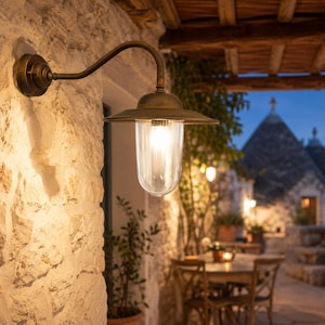 Può includere: Una lanterna da esterno montata a parete con finitura in ottone anticato, dotata di un braccio curvo e un paralume in vetro trasparente a coste. L'apparecchio emette una luce calda e brillante, illuminando la parete in pietra testurizzata.