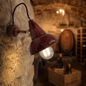 Peut inclure: Une lampe murale de style vintage avec un abat-jour conique bordeaux et une ampoule Edison visible. La lampe est fixée à un mur en pierre avec un câble noir. L'arrière-plan suggère un cadre de cave à vin.