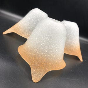 Könnte beinhalten: Drei mattierte Glaslampenschirme mit einem Farbverlauf von Weiß zu hellem Pfirsich. Die Lampenschirme haben eine strukturierte, gesprenkelte Oberfläche.
