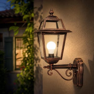 Può includere: Una lanterna da parete per esterni con finitura bronzo. La lanterna presenta un design geometrico con pannelli in vetro trasparente e una lampadina accesa all'interno. Fissata a una staffa decorativa su un muro beige.