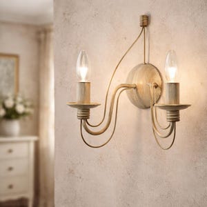 Può includere: Applique da parete color crema con due lampadine a forma di candela. L'applique ha un design a braccio curvo decorativo e una piastra rotonda. I corpi illuminanti sono illuminati, proiettando un bagliore caldo. L'applique è montata su una parete strutturata.
