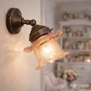 Può includere: Applique da parete in stile vintage con finitura bronzo. La lampada ha un paralume in vetro rosa a forma di fiore. La luce è accesa, emettendo un bagliore caldo. La lampada è montata a parete.
