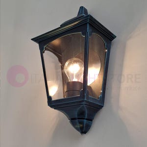 Könnte beinhalten: Eine schwarze und grüne Außenwandleuchte mit einem klaren Glasschirm und einem traditionellen Design. Das Licht ist eingeschaltet und die Glühbirne ist sichtbar.