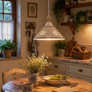 Puede incluir: Escena de cocina rústica con una lámpara colgante blanca con recortes florales, sobre una mesa redonda de madera. La mesa está puesta con un jarrón de flores, una taza y un cuenco de fruta verde. La habitación tiene una estética rústica.
