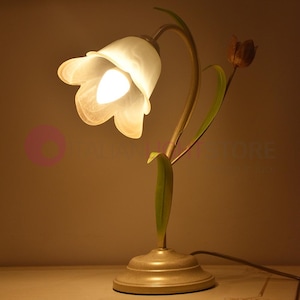 Op de afbeelding: Een tafellamp in de vorm van een bloem met een witte glazen kap en een gouden metalen voet. De lamp heeft een groene steel en bladeren en een bruine bloemknop.