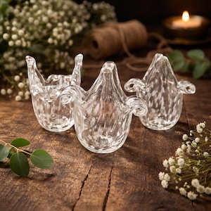 Op de afbeelding: Drie heldere glazen vazen met een gespikkeld patroon. Elke vaas heeft een unieke vorm met uitlopende toppen en decoratieve handvatten. De vazen zijn gerangschikt op een houten oppervlak, met groen en een kaars op de achtergrond.