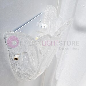 May include: Clear glass wall sconce with a textured, organic design. The sconce is mounted on a white metal plate and features a bulb socket. The text "ITALIANLIGHTSTORE Design in luce" is visible.