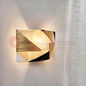 K&ouml;nnte beinhalten: Geometrische Wandleuchte mit Goldblatt- und schwarzem Design. Die Leuchte hat eine rechteckige Form mit einer Reihe geometrischer Formen. Die Leuchte ist an einer wei&szlig;en Wand montiert. Die Leuchte hat ein warmes Licht.