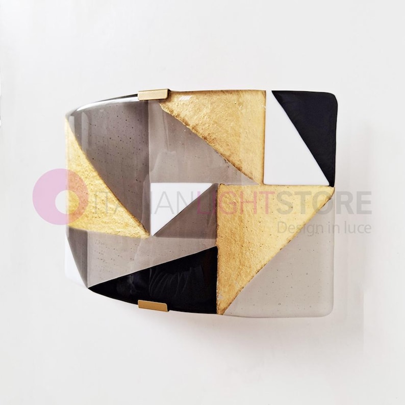 K&ouml;nnte beinhalten: Geometrische Wandleuchte mit goldenen, schwarzen, wei&szlig;en und grauen dreieckigen Akzenten. Die Leuchte hat eine rechteckige Form mit gew&ouml;lbter Ober- und Unterseite. Der Text "AN LIGHTSTORE Design in luce" ist sichtbar.
