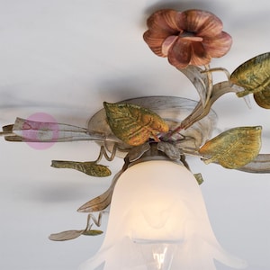 K&ouml;nnte beinhalten: Eine wei&szlig;e Deckenleuchte mit floralem Design. Die Leuchte hat einen wei&szlig;en Glasschirm und Metallbl&auml;tter und -blumen in Gr&uuml;n-, Gelb- und Rosat&ouml;nen.