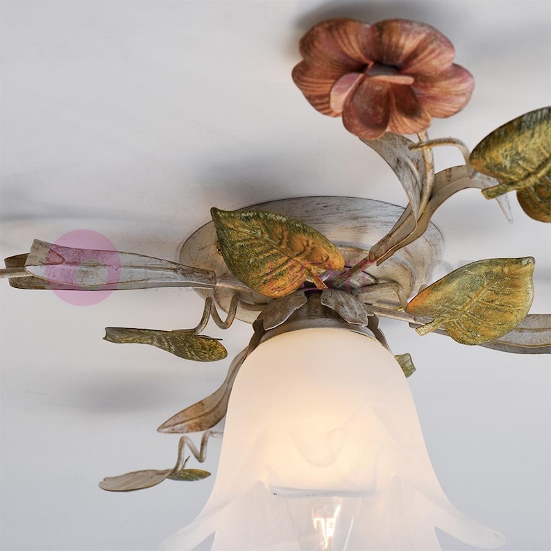 K&ouml;nnte beinhalten: Eine wei&szlig;e Deckenleuchte mit floralem Design. Die Leuchte hat einen wei&szlig;en Glasschirm und Metallbl&auml;tter und -blumen in Gr&uuml;n-, Gelb- und Rosat&ouml;nen.