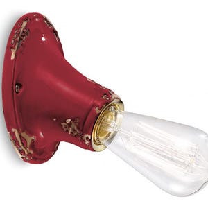 Puede incluir: Una lámpara de pared de cerámica roja con un acabado desgastado y una bombilla de vidrio transparente. La lámpara tiene una forma redondeada y una base de metal dorado.