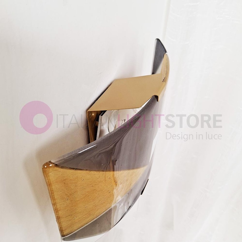K&ouml;nnte beinhalten: Moderne Wandleuchte mit einzigartigem Design. Die Leuchte hat eine geschwungene, transparente Front mit braunen, grauen und hellen Holzfarben. Eine rechteckige, goldfarbene Metallhalterung tr&auml;gt die Leuchte, die an einer wei&szlig;en Wand befestigt ist.