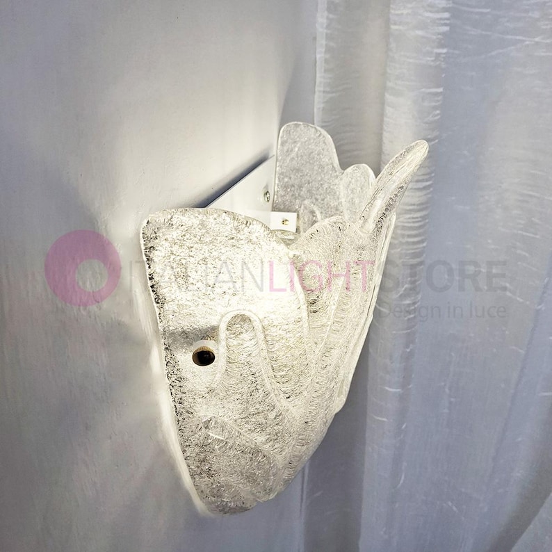 May include: A clear, textured glass wall sconce with a unique, organic shape. The sconce is mounted on a white metal bracket and emits a soft, warm light. The design features a textured surface, creating a play of light and shadow. The sconce is a decorative lighting fixture.