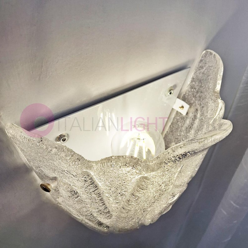 May include: A clear, textured glass wall sconce with a seashell design. The sconce is mounted on a white metal plate and holds a light bulb. The text "ITALIANLIGHT" is visible.