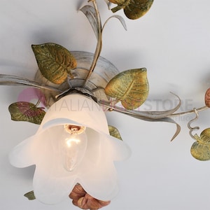 K&ouml;nnte beinhalten: Eine wei&szlig;e Deckenleuchte mit floralem Design. Die Leuchte hat einen wei&szlig;en, mattierten Glasschirm in Form einer Blume. Die Bl&auml;tter sind in Gr&uuml;n- und Braunt&ouml;nen bemalt.