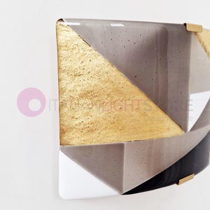 K&ouml;nnte beinhalten: Geometrische Wandkunst mit einem goldenen, grauen und schwarzen Design. Das Kunstwerk hat ein strukturiertes goldenes Dreieck und ist an einer wei&szlig;en Wand befestigt. Das St&uuml;ck hat eine moderne, abstrakte &Auml;sthetik mit Messingakzenten.