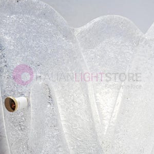 May include: A close-up of a clear, textured glass light fixture. The fixture has a wavy, organic shape and a frosted appearance. A brass and white light socket is visible on the left side. The text "ITALIANLIGHTSTORE Design in luce" is visible.