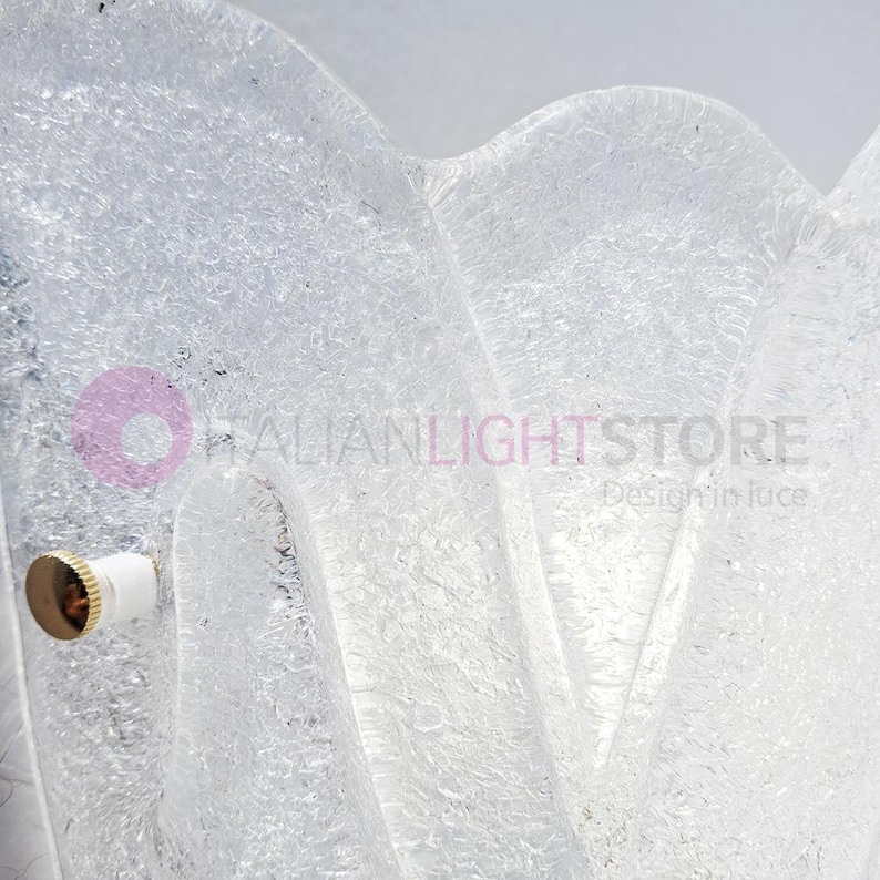 May include: A close-up of a clear, textured glass light fixture. The fixture has a wavy, organic shape and a frosted appearance. A brass and white light socket is visible on the left side. The text "ITALIANLIGHTSTORE Design in luce" is visible.