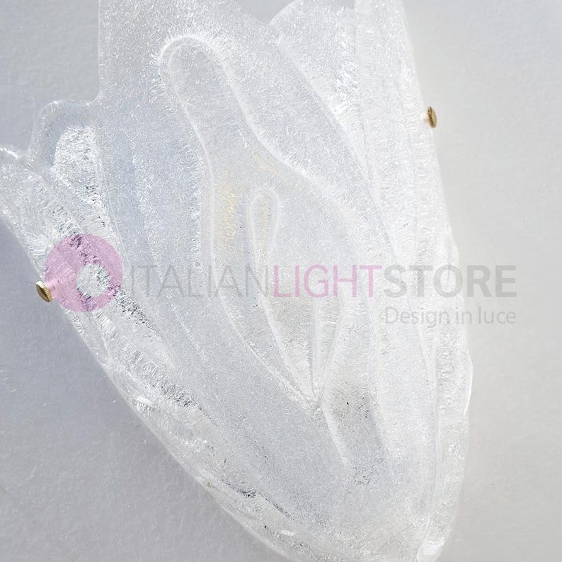 May include: A clear, textured glass wall sconce with a floral design. The sconce is mounted on a white wall and has brass-colored hardware. The text "ITALIANLIGHTSTORE Design in luce" is visible.