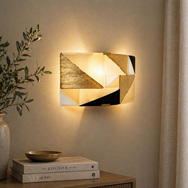 K&ouml;nnte beinhalten: Geometrische Wandleuchte mit goldenem, schwarzem und wei&szlig;em Design. Die Leuchte ist rechteckig mit einem Patchwork aus Formen und einem warmen Schein. Die Leuchte ist an einer beigefarbenen Wand montiert, darunter B&uuml;cher und eine dekorative Schale.