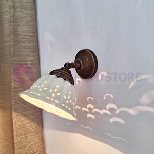 Puede incluir: Aplique de pared de cerámica blanca con un patrón decorativo de corte. La aplique tiene un acabado en bronce y está montada en una pared blanca.