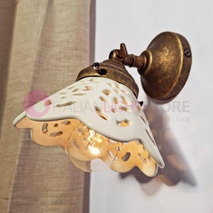 Può includere: Un'applique da parete in ceramica bianca con un design a taglio decorativo. L'applique ha una finitura in ottone ed è montata su una parete. L'applique è illuminata da una lampadina.