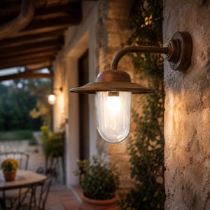 Può includere: Una lampada da parete per esterni con finitura in bronzo scuro, dotata di un braccio curvo, un ampio paralume circolare e un diffusore cilindrico in vetro trasparente a coste verticali che racchiude una lampadina accesa su una parete in pietra.