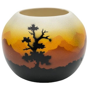 Peut inclure: Un vase rond en céramique avec un motif de paysage. Le vase présente un dégradé de couleurs, allant du crème en haut au noir à la base. Une silhouette noire d'arbre est centrée sur le vase, avec des formes de montagnes orange et marron.
