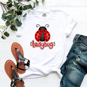 Può includere: Maglietta bianca con un disegno di coccinella rossa e nera e la scritta "ladybug". L'immagine mostra anche jeans blu e sandali marroni.