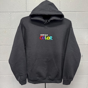 Puede incluir: Una sudadera negra con capucha y manga larga que presenta un bolsillo tipo canguro frontal y un logotipo bordado en el pecho. El logotipo muestra el texto GAME BOY en blanco, con la palabra COLOR debajo en letras multicolores, infladas y redondeadas.