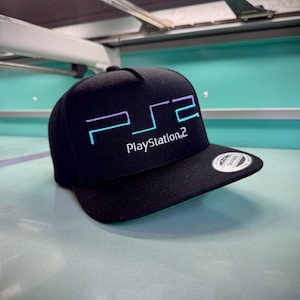 May include: A black snapback cap featuring an embroidered PlayStation 2 logo on the front. The logo displays the letters PS2 in a gradient of light blue to purple, with the text PlayStation 2 written in white underneath. A circular sticker is on the brim.