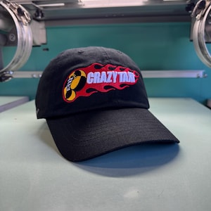 May include: A black baseball cap featuring an embroidered logo on the front. The logo displays the text CRAZY TAXI in white letters, surrounded by a stylized red and yellow flame design. The cap has a curved brim and a structured crown.