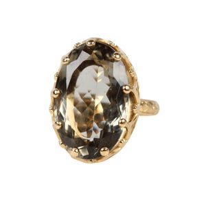 May include: A gold ring featuring a large, oval-cut, translucent smoky quartz gemstone. The stone is held in a decorative, scalloped gold setting with small, rounded prongs along the edge, attached to a textured, thin gold band.