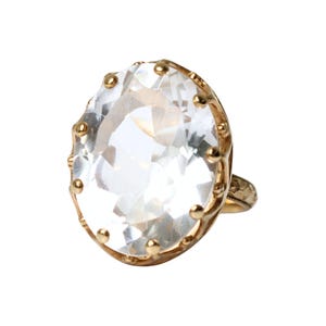 May include: A gold ring featuring a large, oval-cut clear gemstone set in a prong setting. The metal band displays an intricate, engraved floral pattern along its surface, providing a detailed texture that complements the faceted, transparent stone.