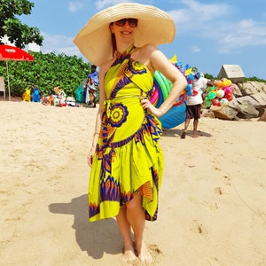 May include: A person on a sandy beach wearing a large straw hat and sunglasses. They are wearing a vibrant yellow wrap dress with a blue and orange pattern. The dress is tied at the waist and has a ruffled hem, creating a flowing silhouette.