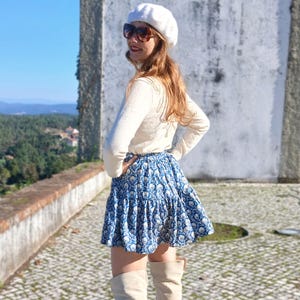 May include: A blue and white floral print skirt with a ruffled hem. The skirt is paired with a cream-coloured long-sleeve shirt and knee-high boots. A white beret and sunglasses complete the outfit. The background features a scenic view.