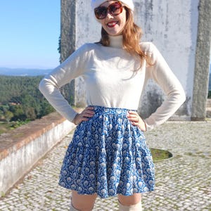 May include: A blue and white floral print skirt. The skirt is worn with a cream-coloured long-sleeved top. The outfit is completed with a white beret and sunglasses. The background features a stone wall and a scenic view.